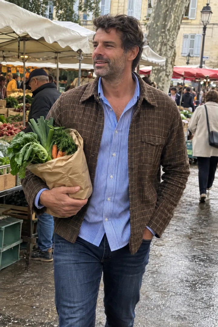 Sur un marché, un homme porte une surchemise à carreaux marron sur une chemise à fines rayures bleues et tient un sac de légumes.