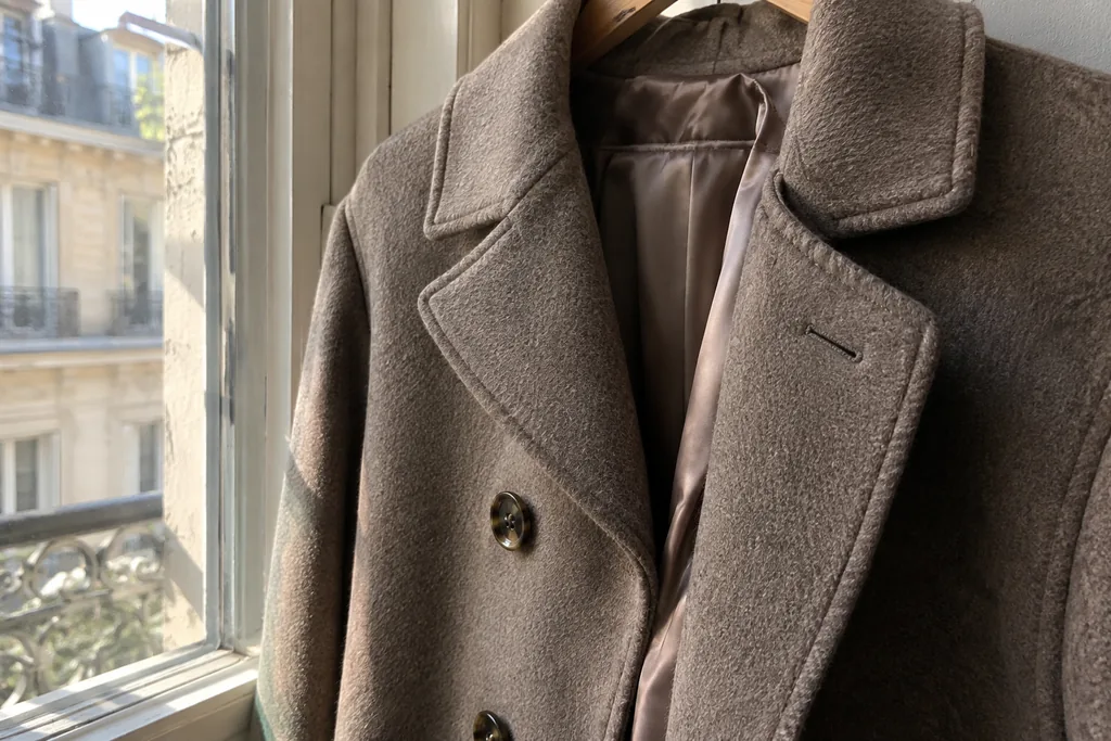 Gros plan sur un manteau en laine suspendu près d’une fenêtre : revers crantés, boutons en corne, surpiqûres nettes et doublure satinée.
