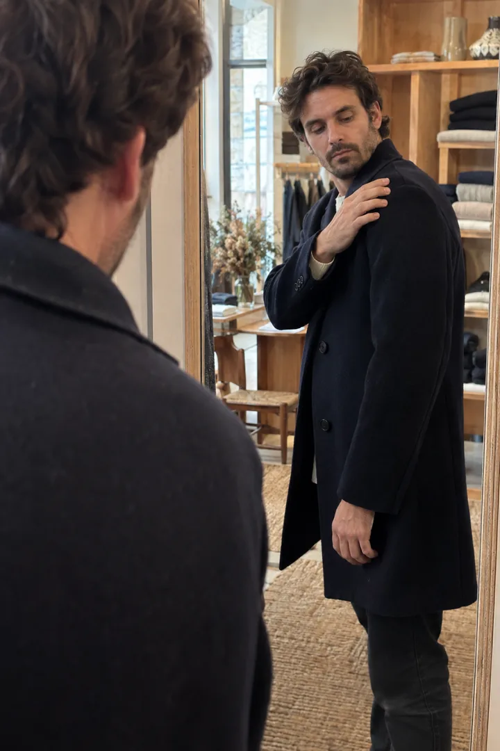Dans une petite boutique, un homme essaie un pardessus marine devant un miroir et vérifie la couture de l’épaule.