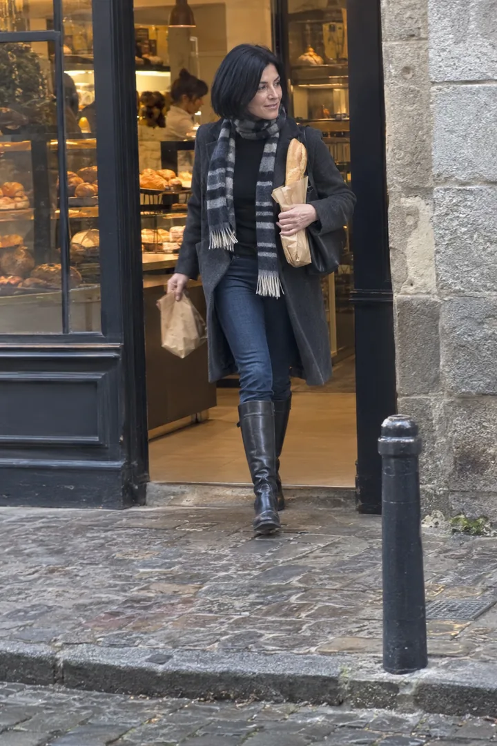 Une femme au carré noir sort d’une boulangerie en jean slim rentré dans des bottes cavalières noires, manteau gris et écharpe rayée, une baguette à la main.
