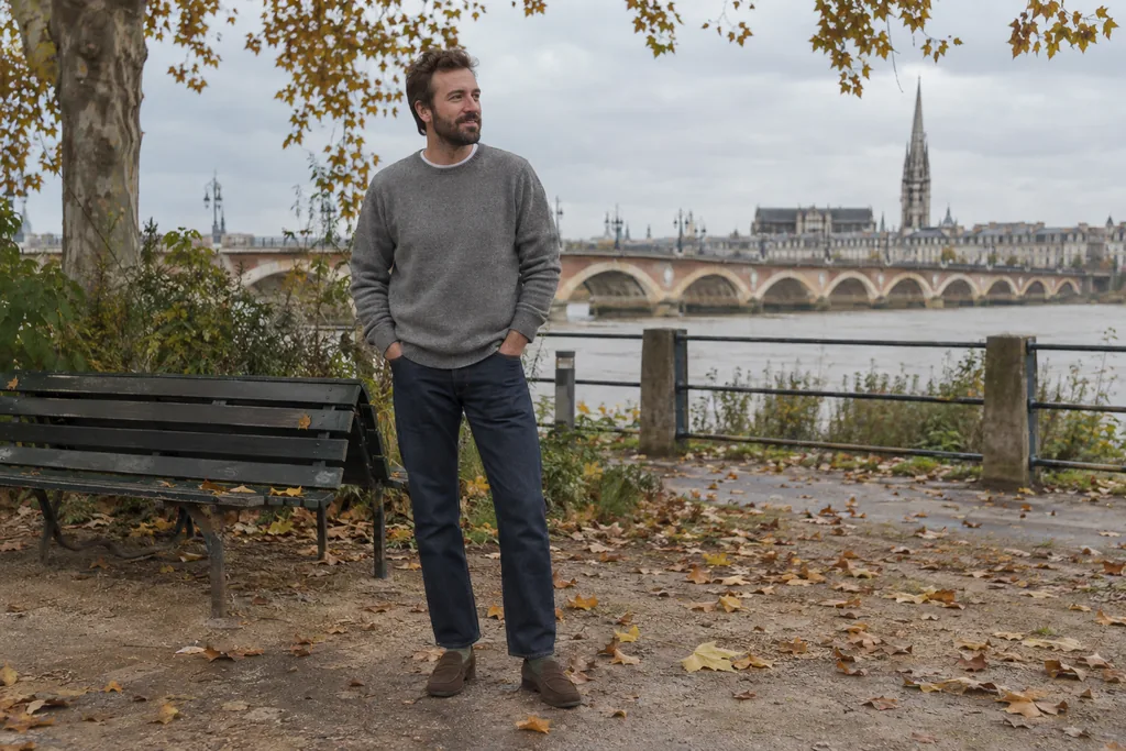 Homme en pull gris et jean indigo portant des chaussettes olive avec des mocassins marron dans un parc.