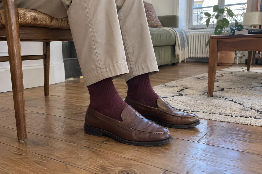 Jambes d’une personne assise en pantalon beige, chaussettes bordeaux et mocassins marron.