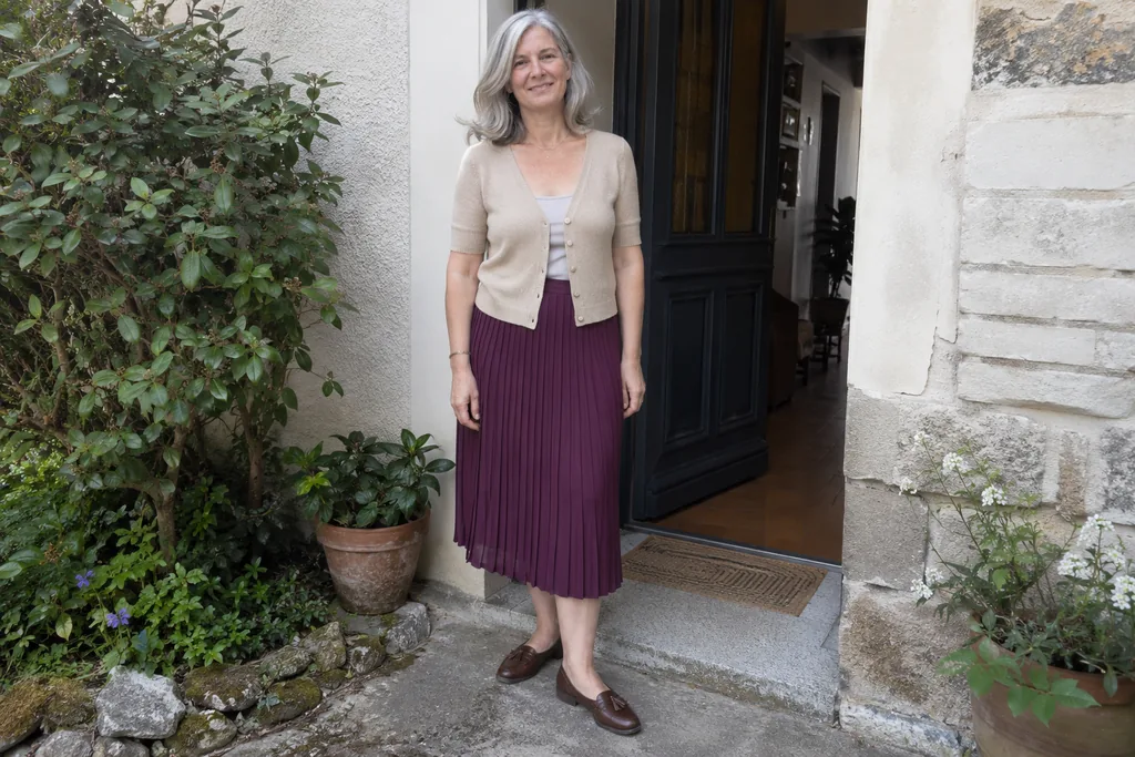Femme aux cheveux gris en cardigan beige, jupe plissée prune et chaussures marron, debout près d’une porte.