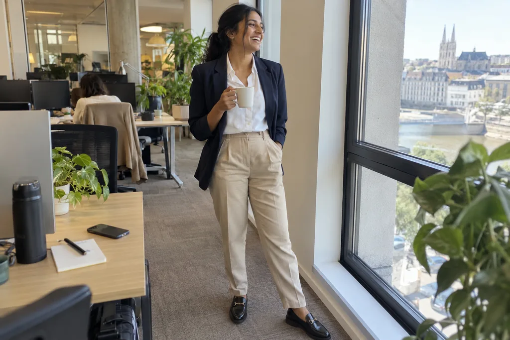Une femme d’une trentaine d’années près d’une fenêtre de bureau en pantalon beige à pinces, chemise blanche, blazer marine et mocassins noirs, une tasse à la main.