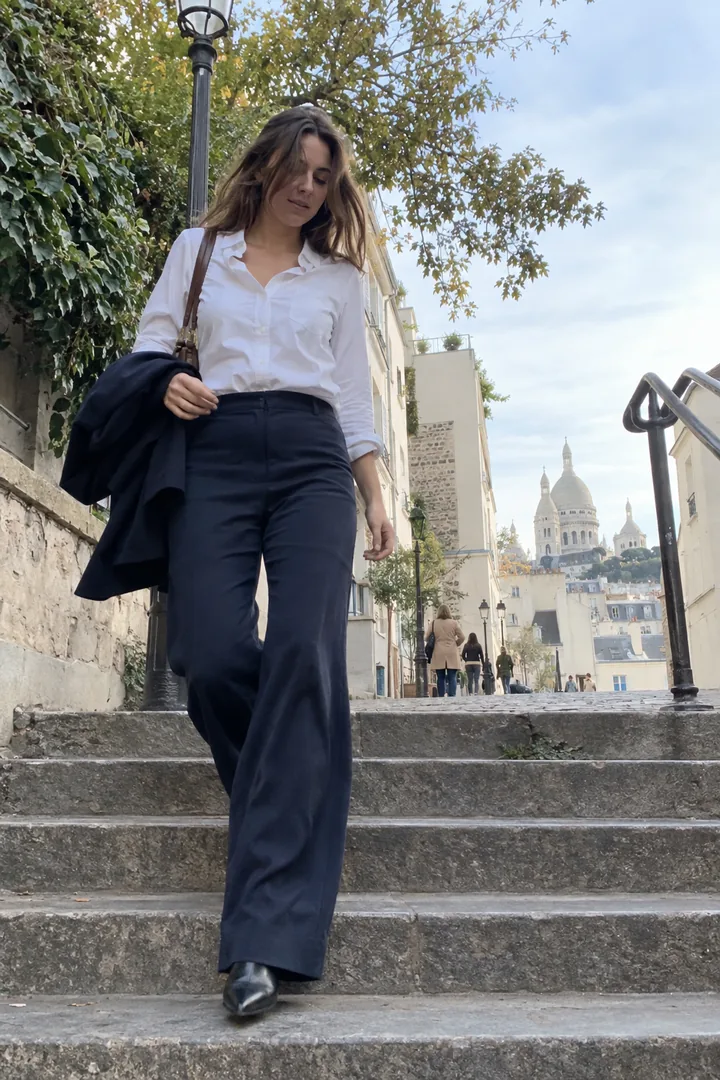 Une femme descend les escaliers de Montmartre en pantalon large marine qui retombe sur des bottines pointues et chemise blanche.