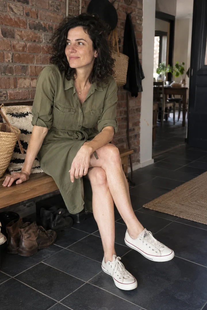 Une femme en robe chemise vert olive et baskets en toile est assise sur un banc.
