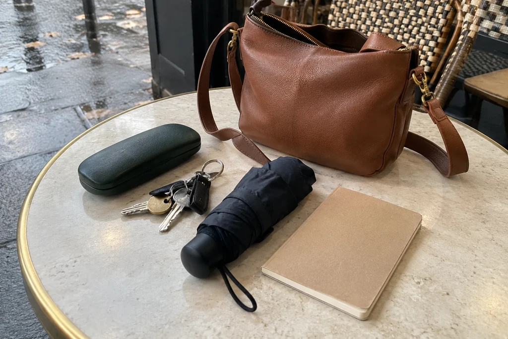 Un étui à lunettes, des clés, un petit parapluie et un carnet sont posés à côté d’un sac sur une table.
