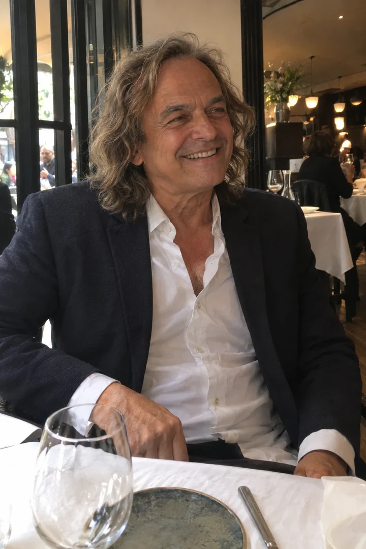 Un homme en veste souple et chemise à col ouvert est assis à une table de restaurant.