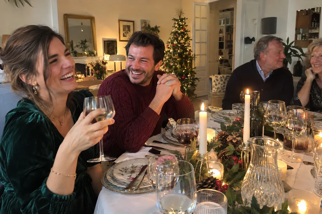 Lors d’un dîner de Noël en famille aux chandelles, une femme en robe de velours vert et un homme en pull bordeaux rient autour de la table.