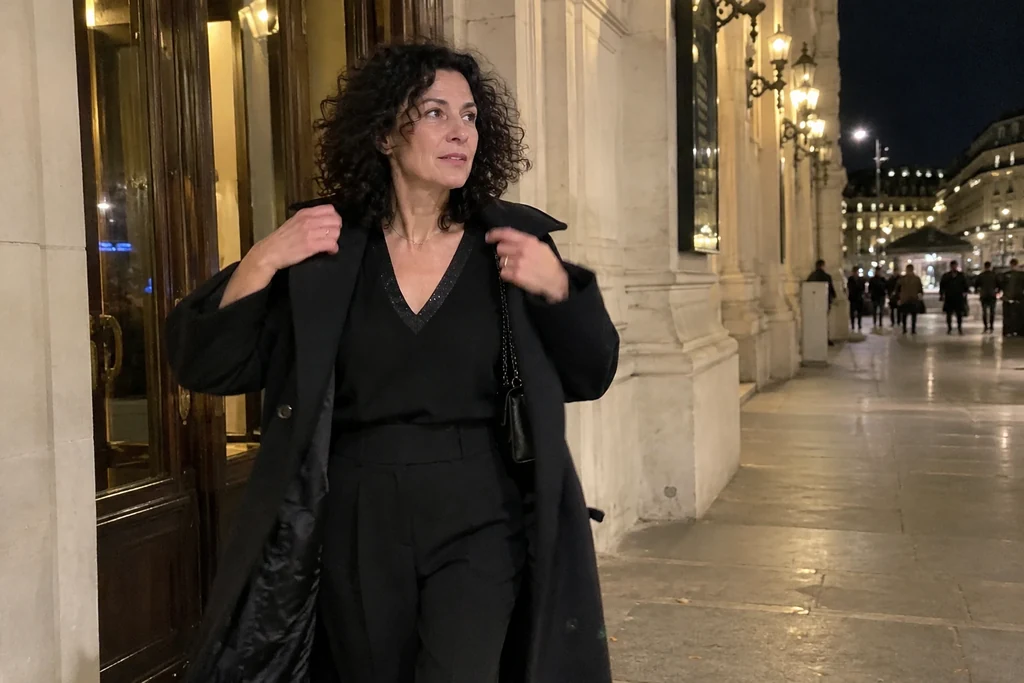 Une femme en tenue de soirée avec pantalon enfile un manteau près d’une porte après la tombée de la nuit.