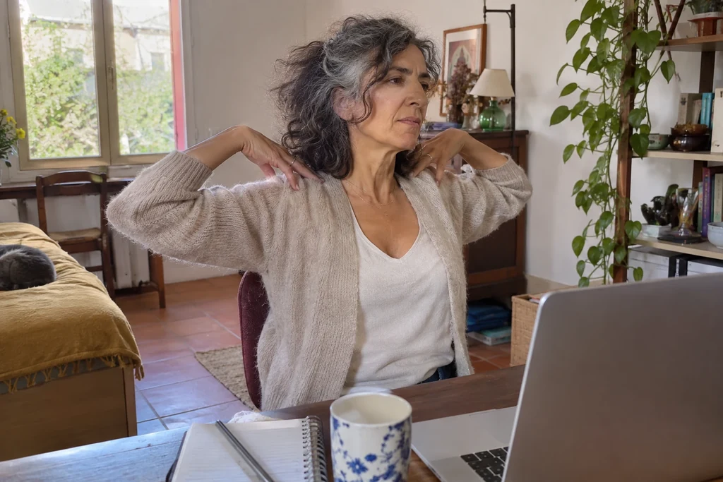 Une femme en cardigan vérifie le mouvement de ses épaules assise à son bureau.