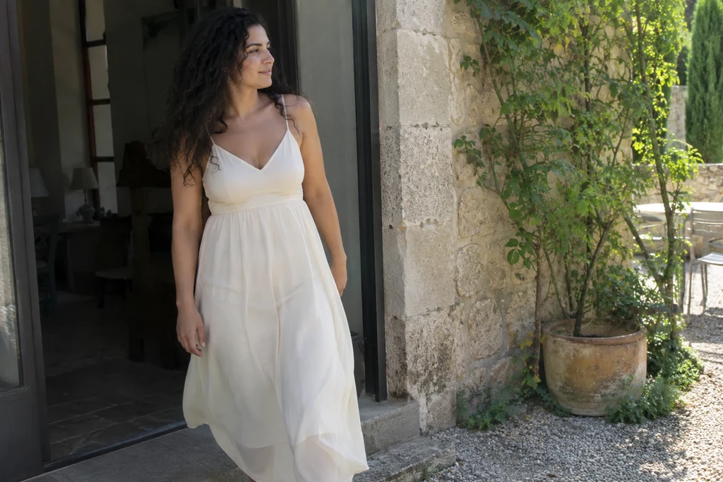 Une femme en robe claire doublée avance dans une cour baignée de lumière naturelle.